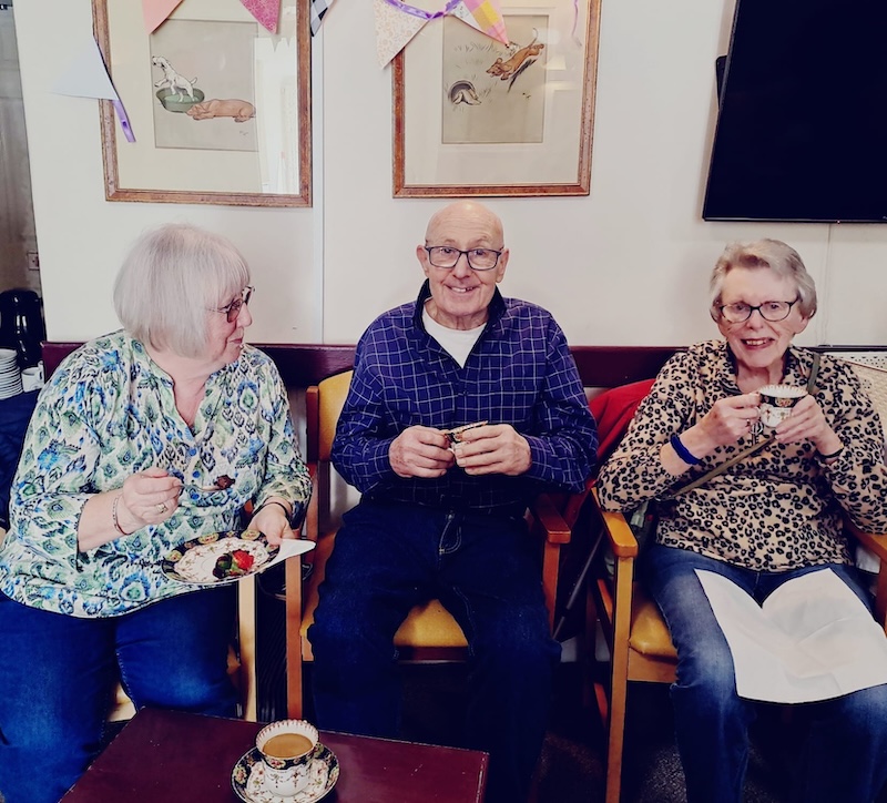 residents sat smiling in the reading room with hot drinks in their hands