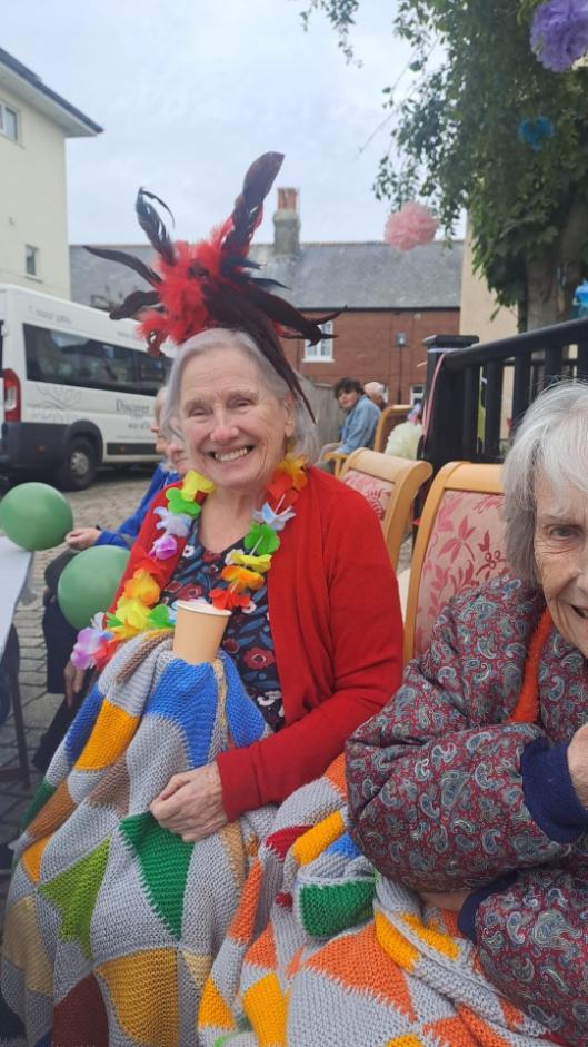 older lady sat down with flowers around her neck and a flowery hat on